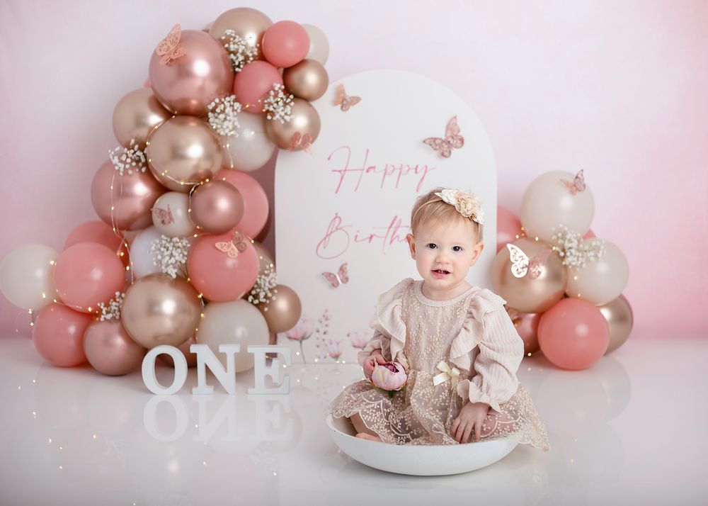 Pink and gold balloon arch with white lettering creates an elegant first birthday party backdrop and photo setup.