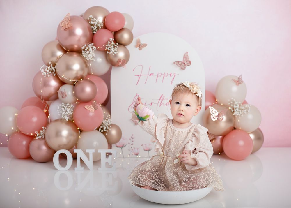 Pink and rose gold balloon arch decorates a delicate first birthday cake smash setup with butterflies and floral accents.