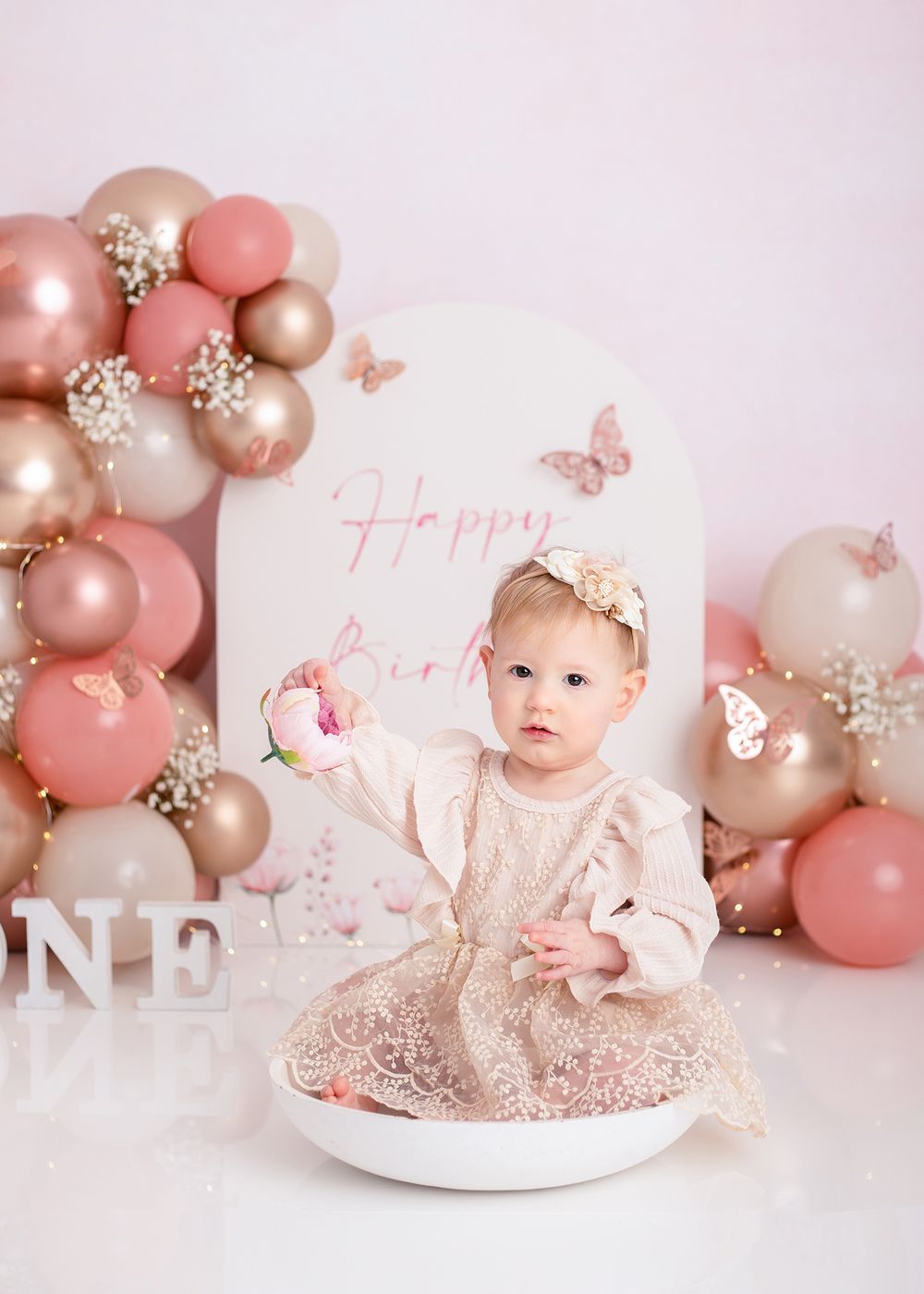 Pink and gold balloon decorations create an elegant first birthday cake smash setup with butterflies and floral accents.