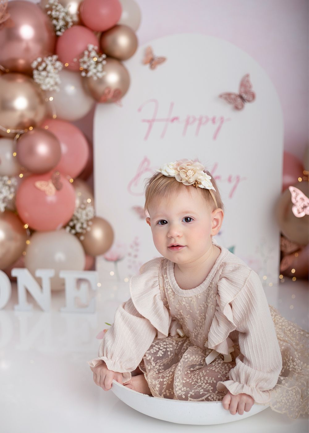 Elegant first birthday celebration setup with pink and gold balloons, cake smash scene, and delicate butterfly decorations.