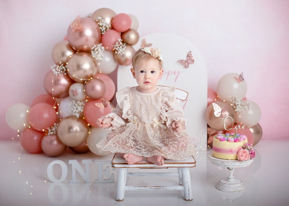 Pink and white balloon arch decorates first birthday celebration setup with white bench and matching cake display.