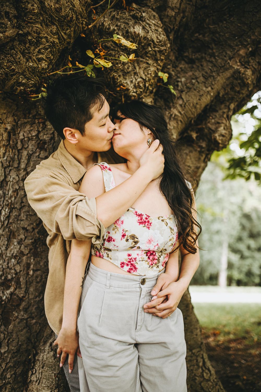 A romantic couple in coordinated outfits sharing intimate moments against a tree in a park setting.