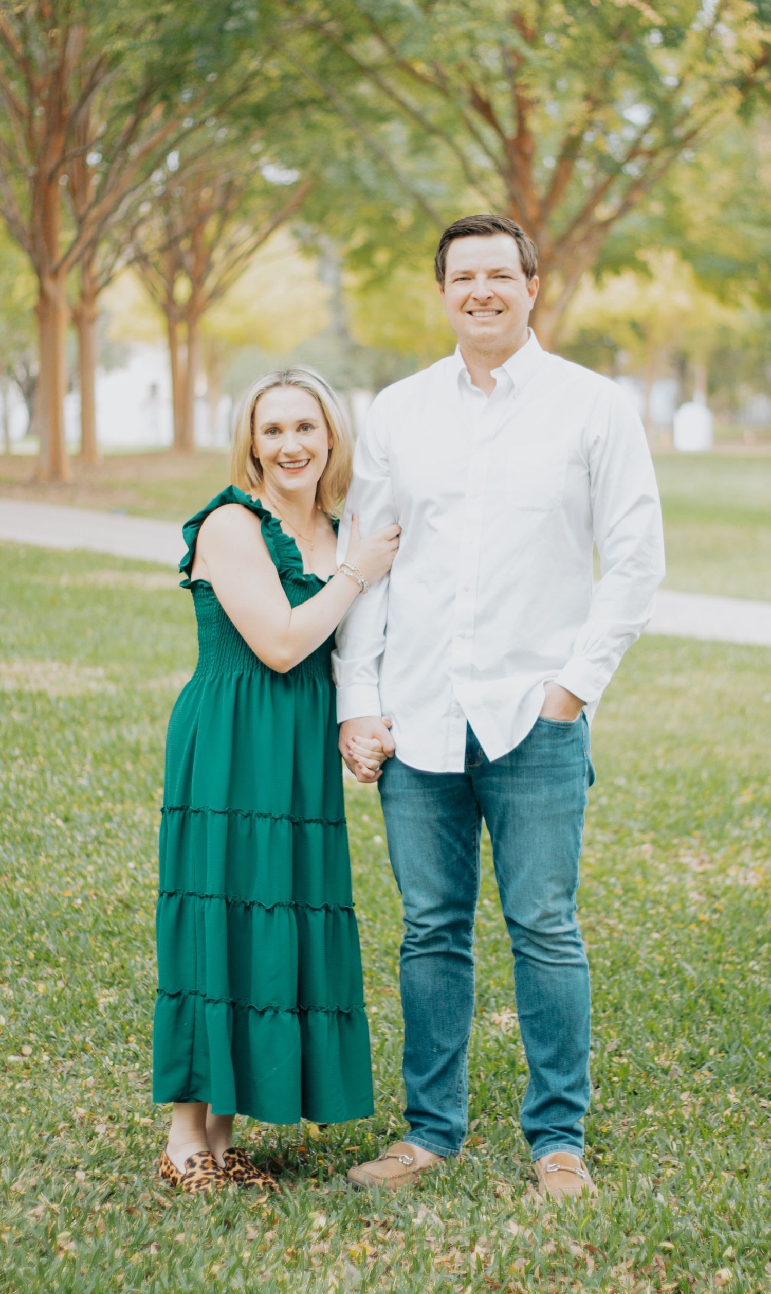 A couple stands together outdoors in a park setting with green trees, wearing coordinated casual attire.
