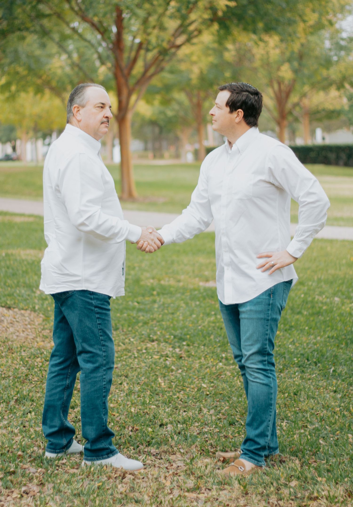 Two people in white shirts and blue pants shake hands in a grassy outdoor setting during autumn.