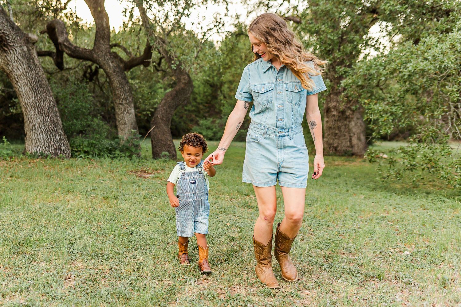 Two people wearing matching denim outfits hold hands while walking together in a grassy area with trees.