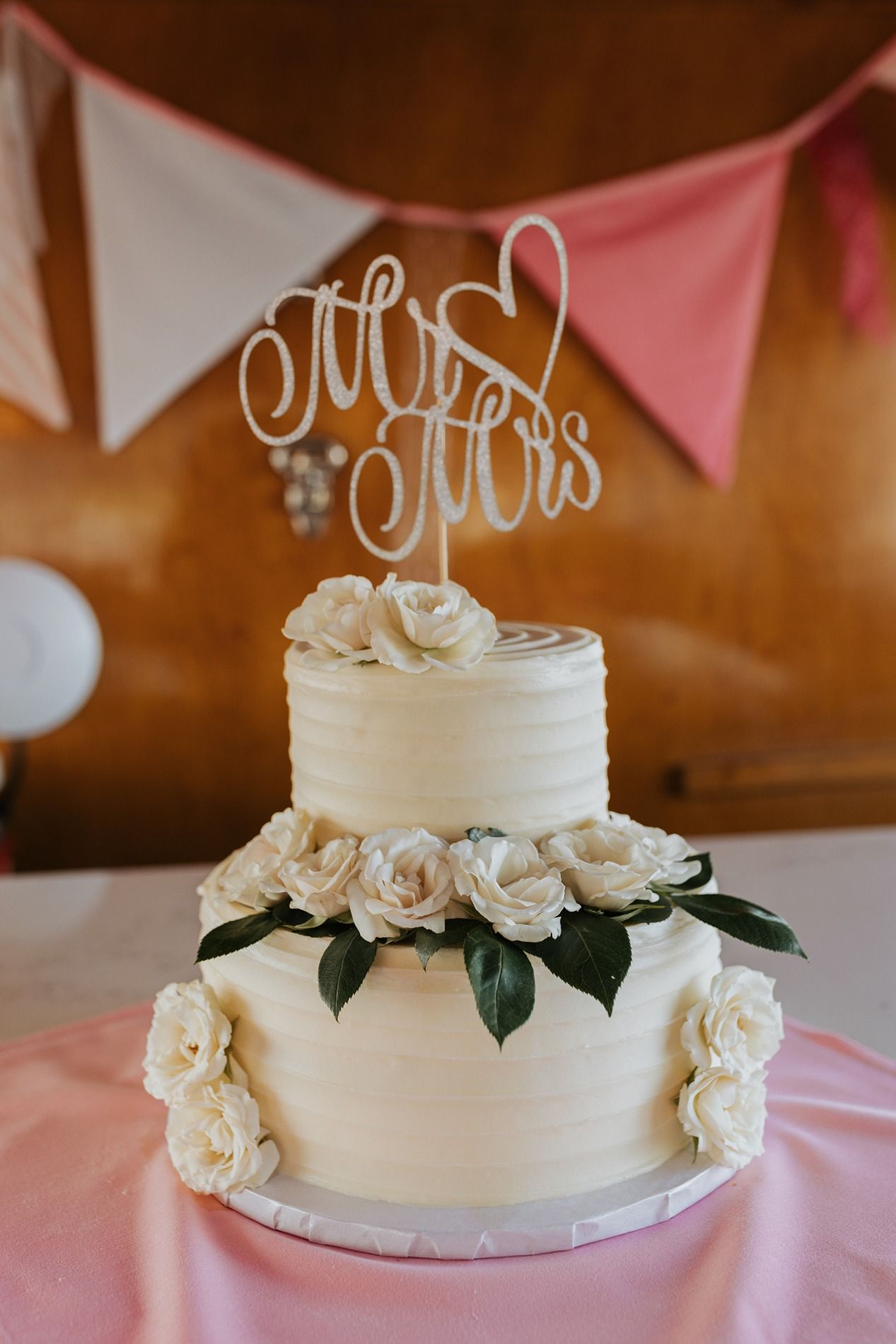 White two-tiered wedding cake decorated with fresh roses and greenery on display at reception with pink and white bunting.