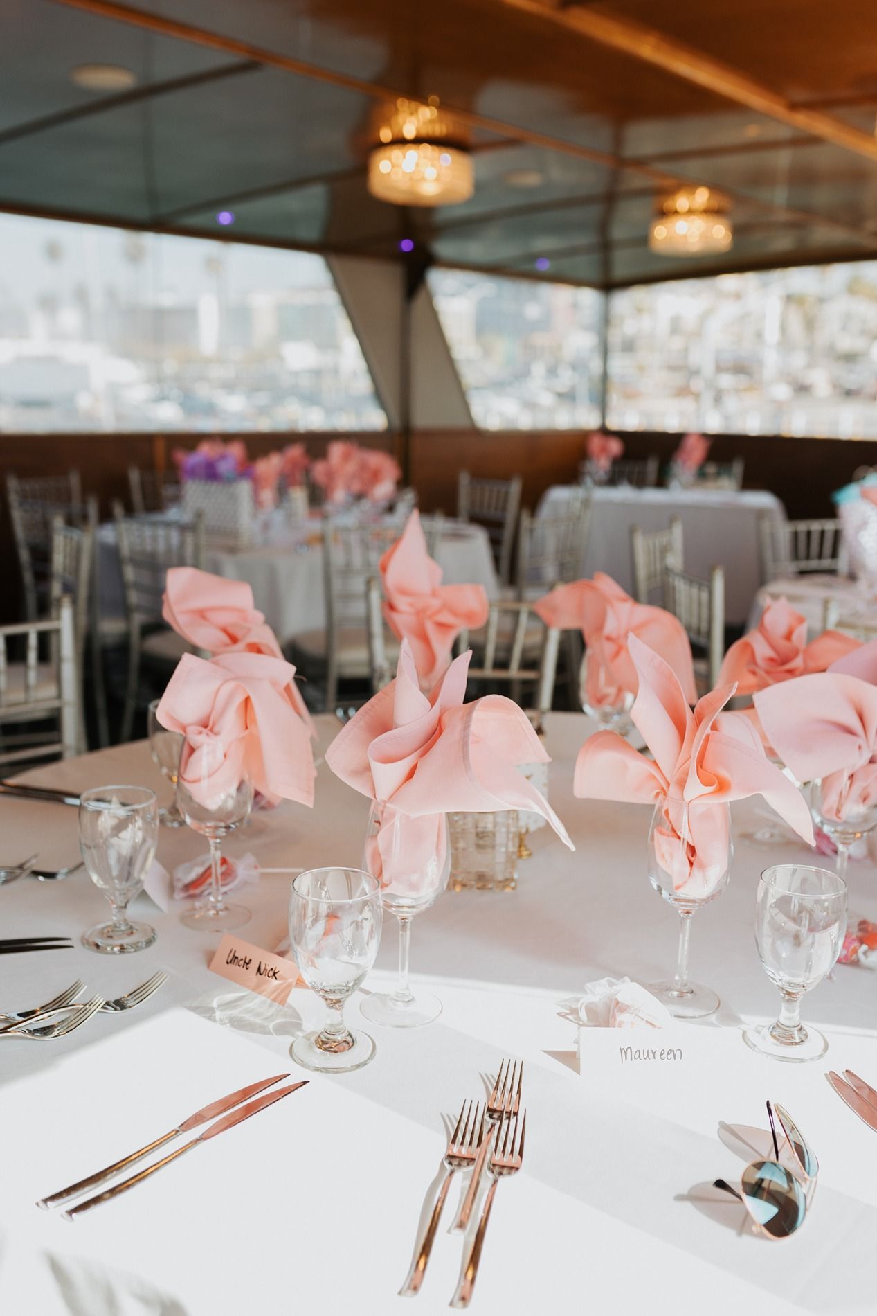 Elegant wedding reception table settings with white linens, pink napkins, and crystal stemware in a formal dining room on a boat.