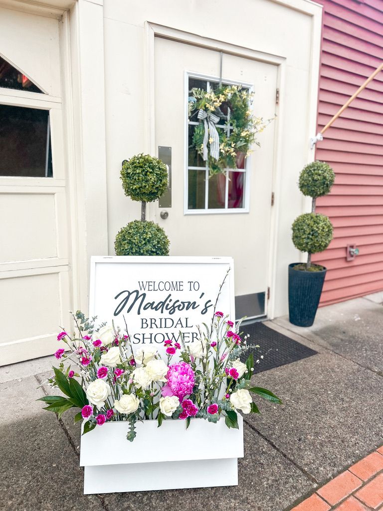 Welcome sign for Madison's Bridal Shower surrounded by pink and white floral arrangement at venue entrance.