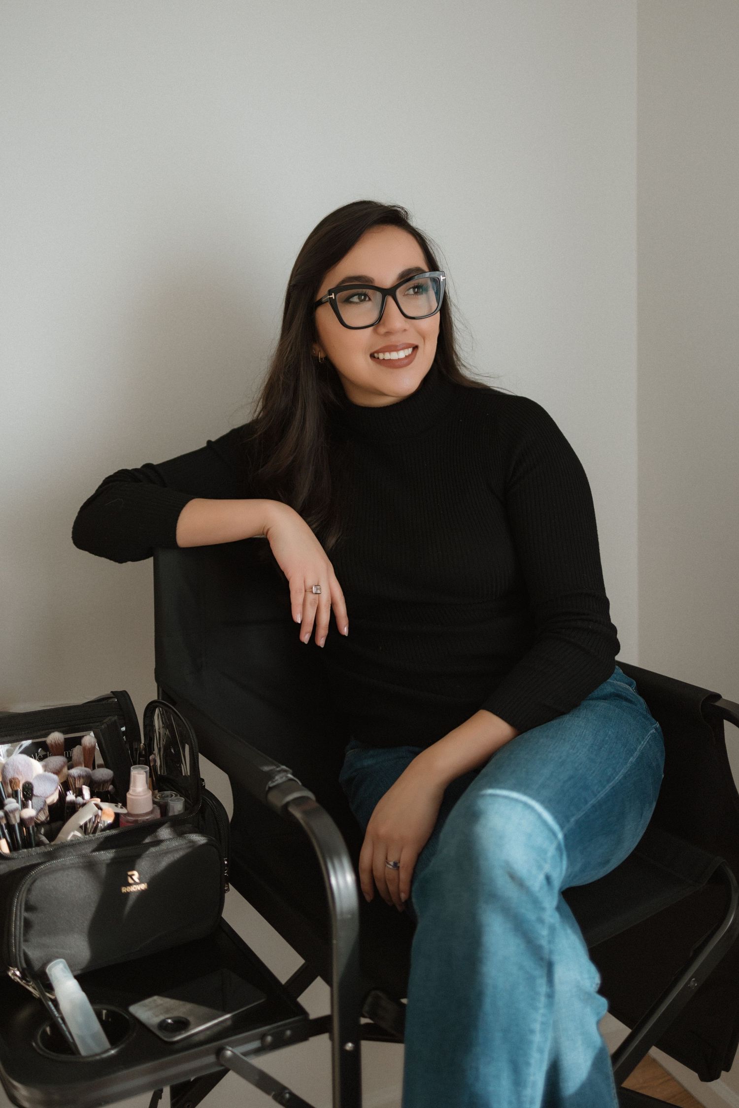 A person in black attire and glasses sits casually on a dark chair in a minimalist setting.