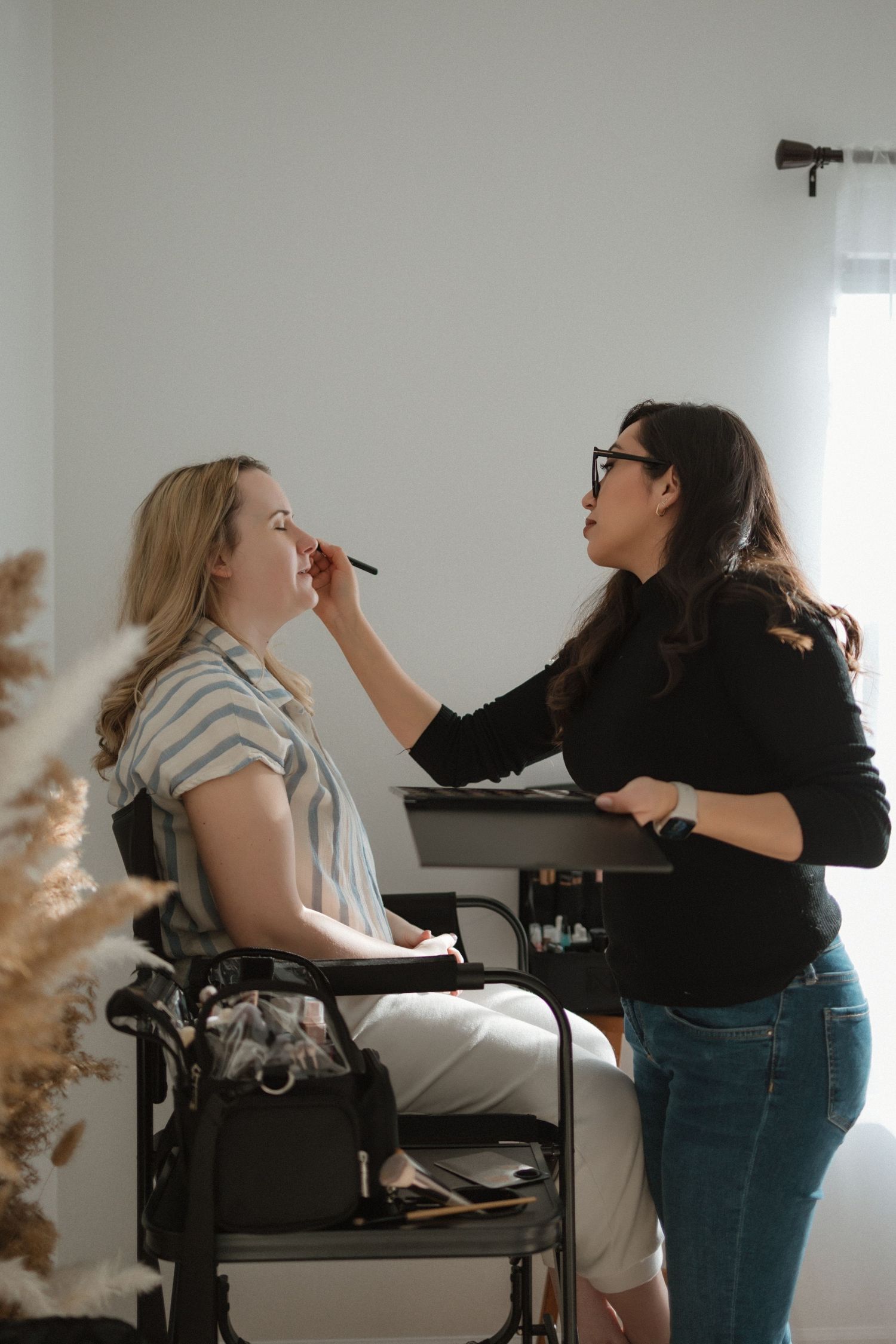 A makeup artist applies cosmetics to a client seated in a chair in a bright white studio space.