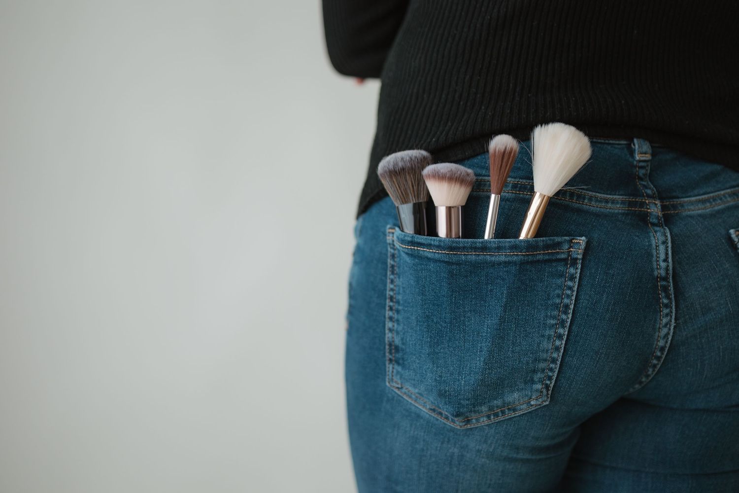 Makeup brushes tucked into back pocket of blue denim jeans against light gray background.