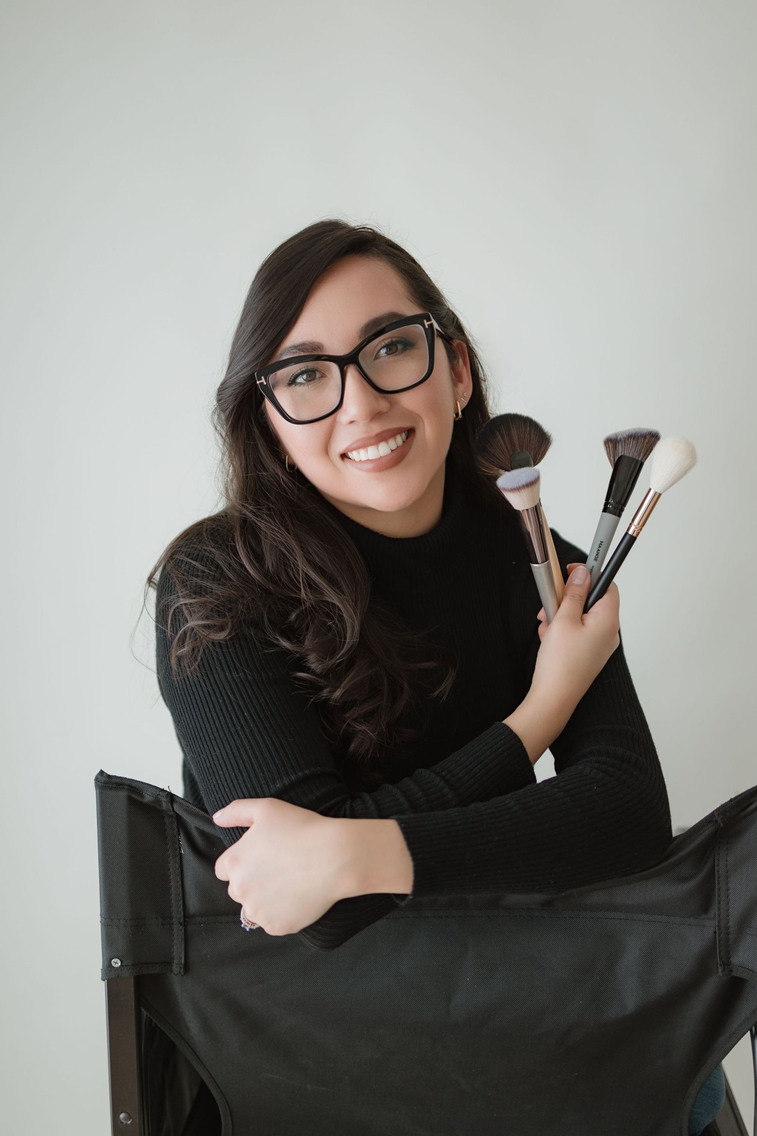 Professional makeup artist holding brushes and posing with black makeup bag against white background.