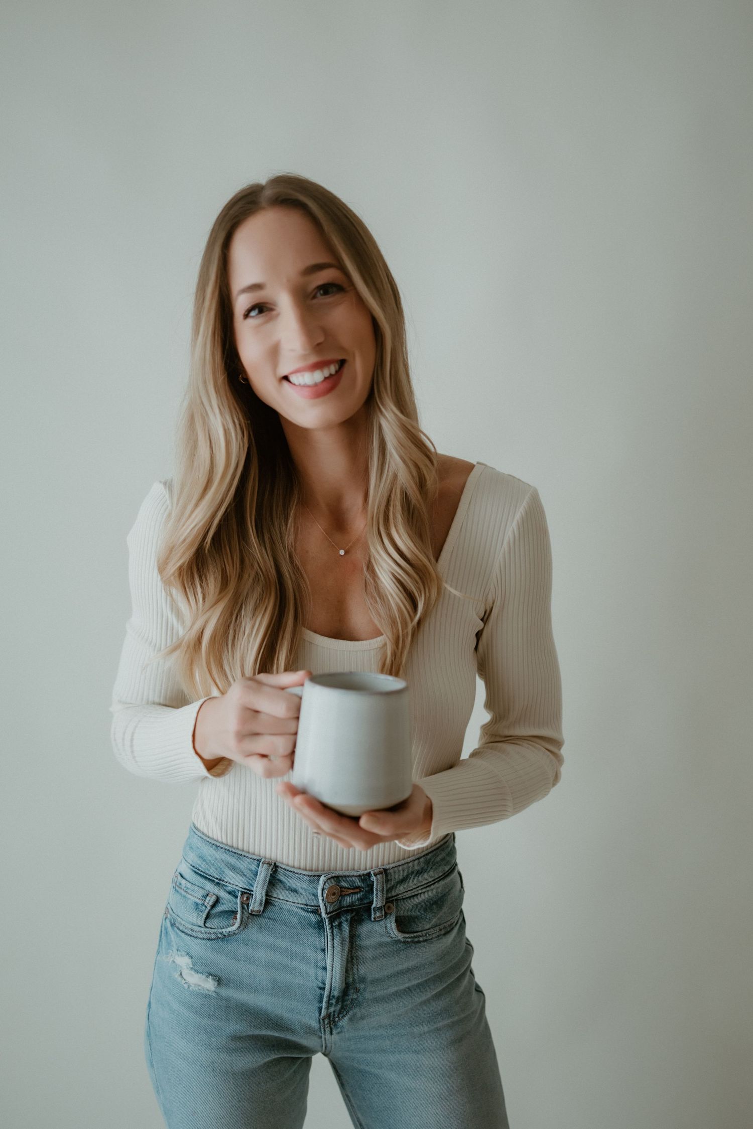 A person in a white sweater and light wash jeans poses casually against a neutral wall in a lifestyle photo series.