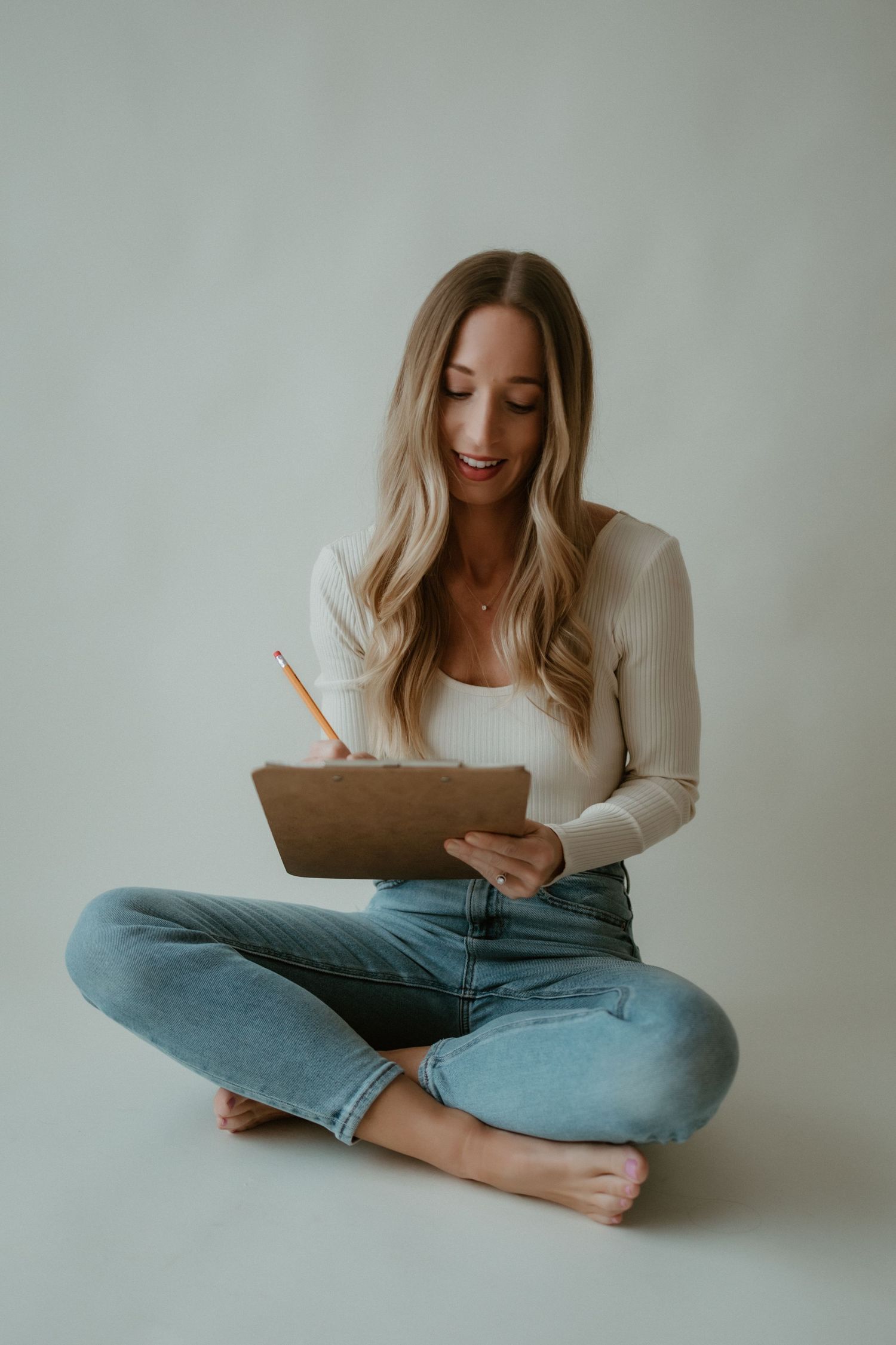 Person in cream sweater and blue jeans sits cross-legged while writing in journal against white background.