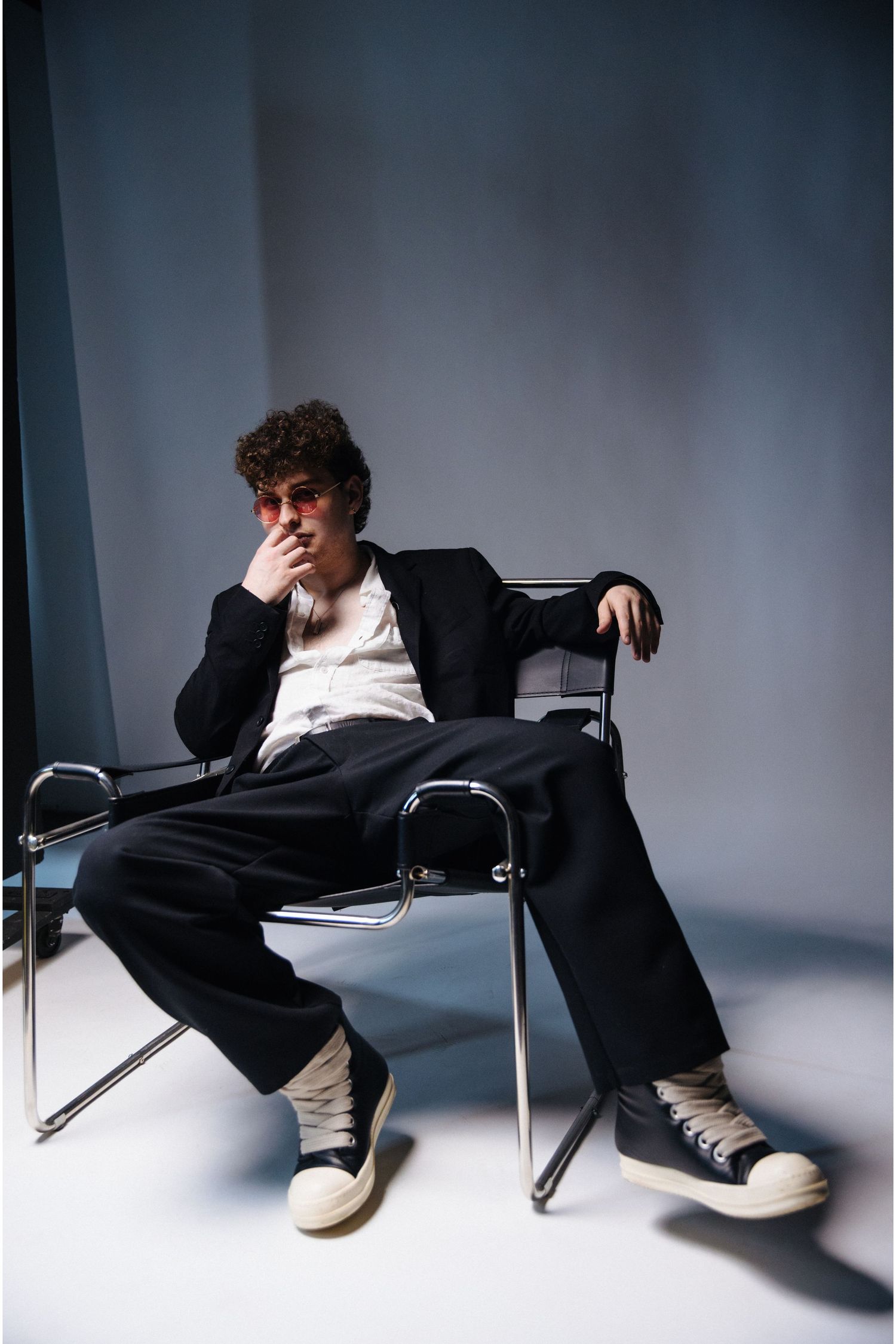 Artistic studio portrait of someone in casual attire with blazer sitting relaxed in a modern chair against white backdrop.