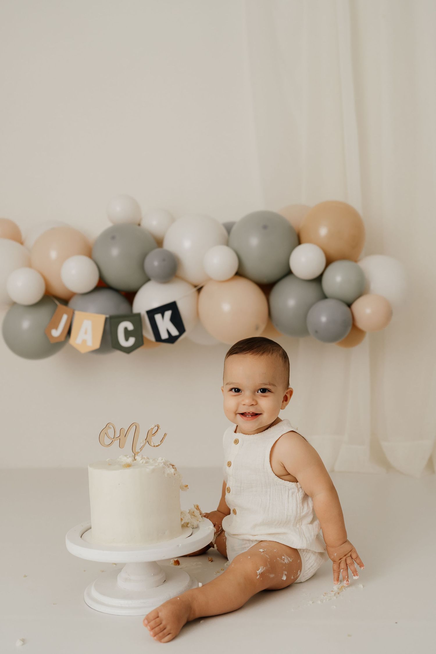 A minimalist cake smash photo setup with grey and white balloons and a simple white cake against a neutral backdrop.