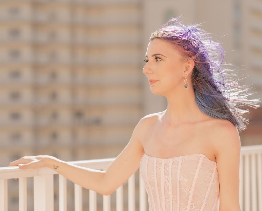 Woman in pink dress poses outdoors against stone wall in bright sunlight with hair blowing in breeze.