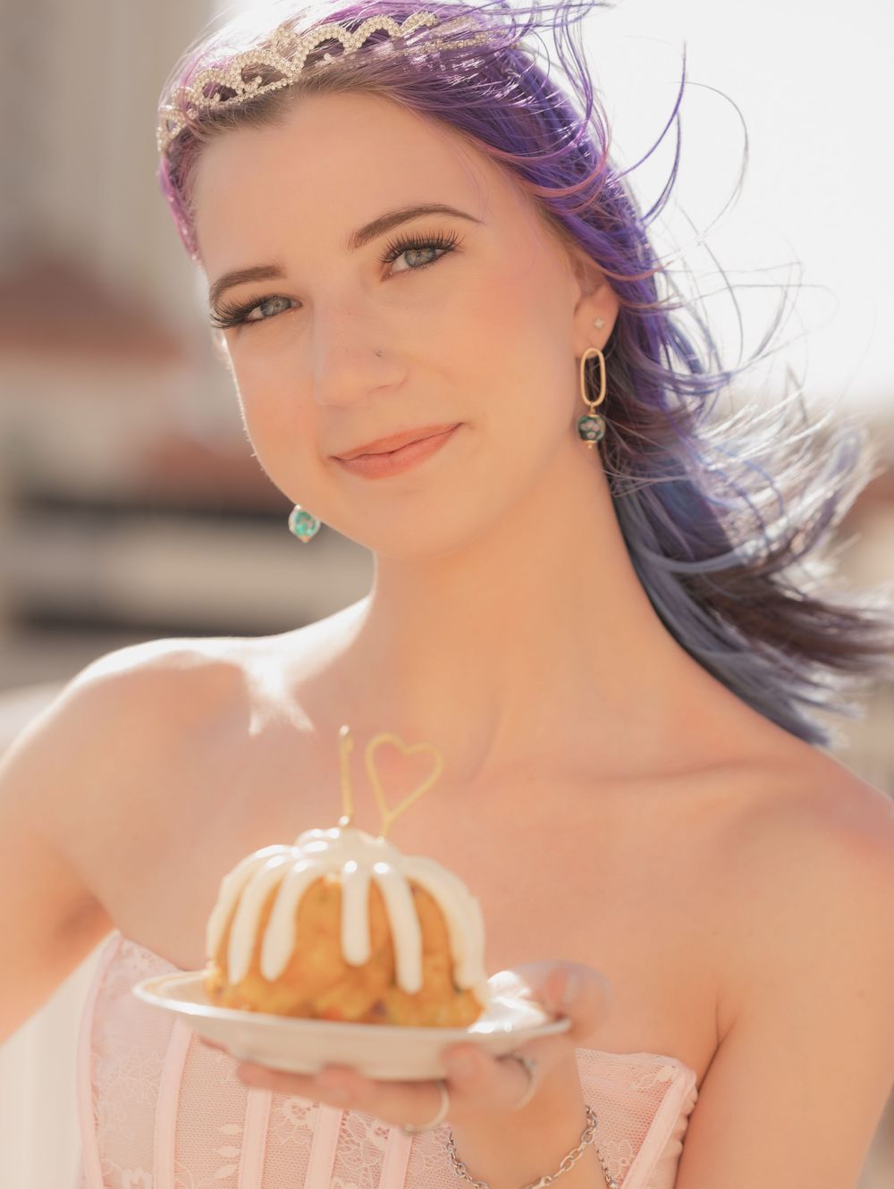 A person with purple hair and a delicate tiara poses in soft sunlight wearing elegant jewelry.