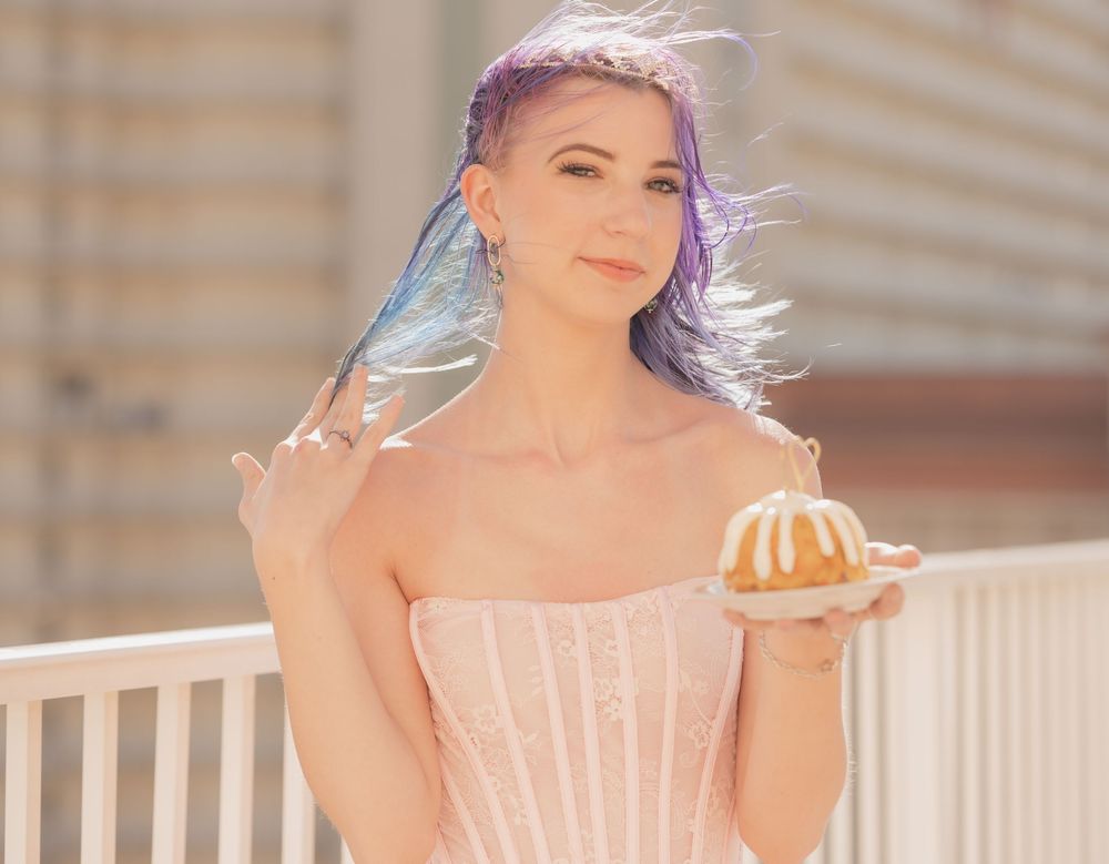 A woman with pastel blue hair in a pink corset dress holds a small bundt cake in sunlit outdoor setting.