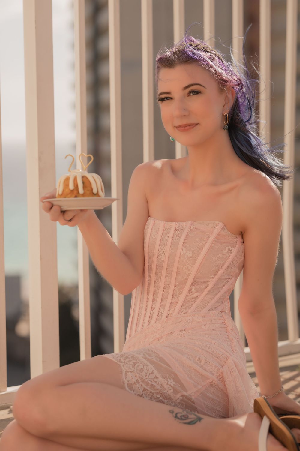 A figure in a pink strapless dress poses with a small cake on a balcony during sunset.