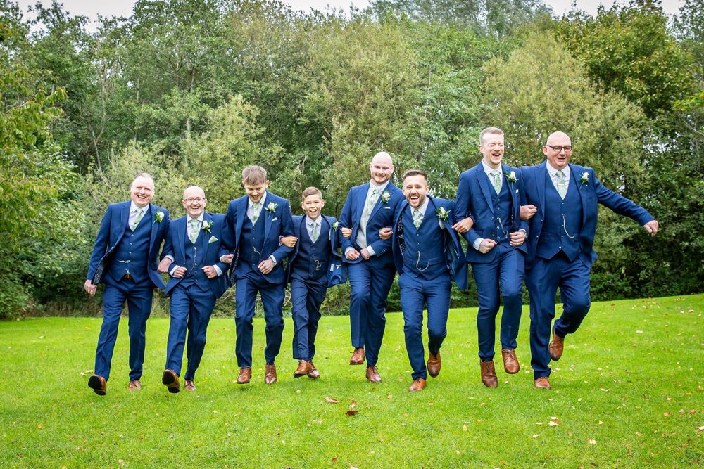 Groom and groomsmen in matching navy suits run and jump playfully across green lawn together.