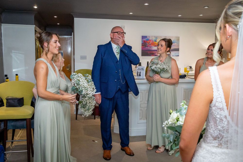 Emotional father wipes tears seeing bride in wedding dress with bridesmaids holding floral bouquets.