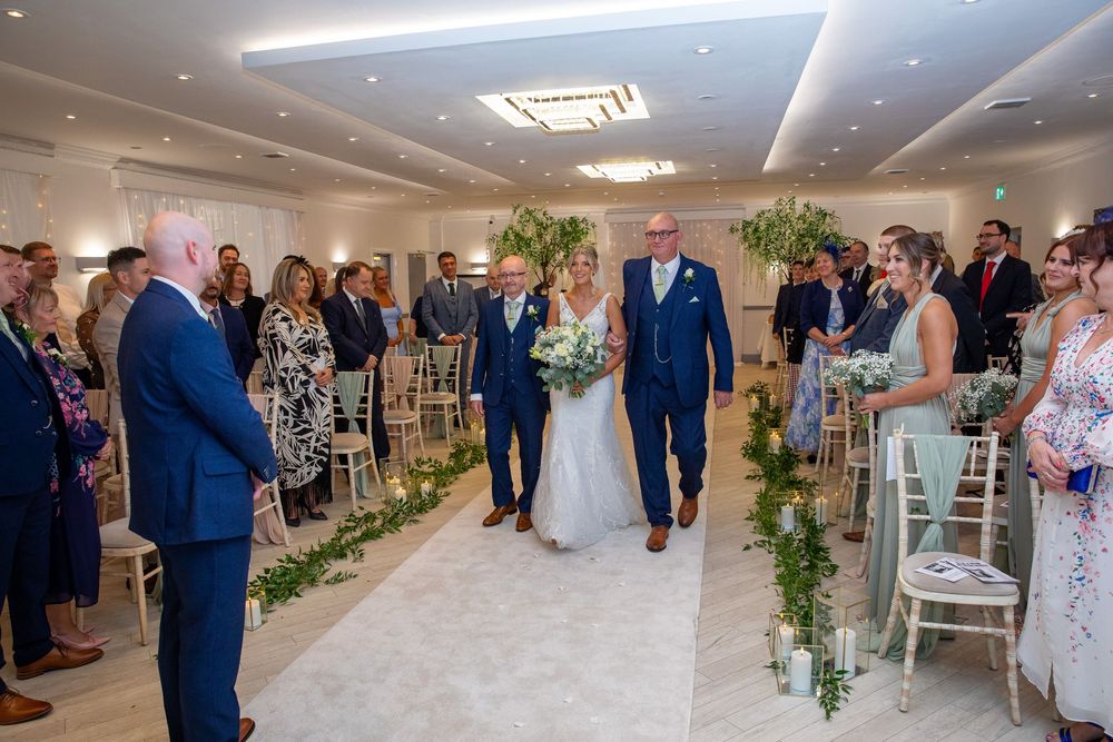 Bride walks down floral aisle escorted by two men toward groom during elegant wedding ceremony.