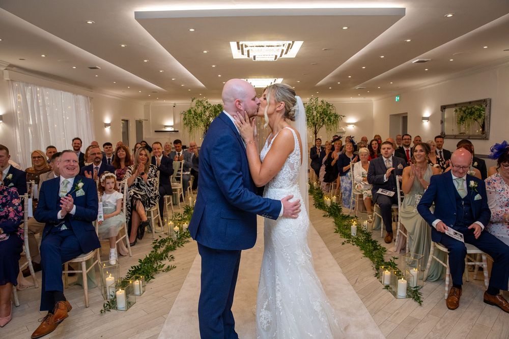 Bride and groom share first kiss at wedding ceremony altar with guests seated in elegant venue.