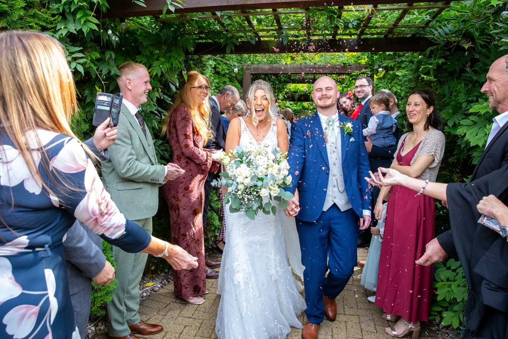 Joyful bride and groom walk through garden archway surrounded by cheering wedding guests.