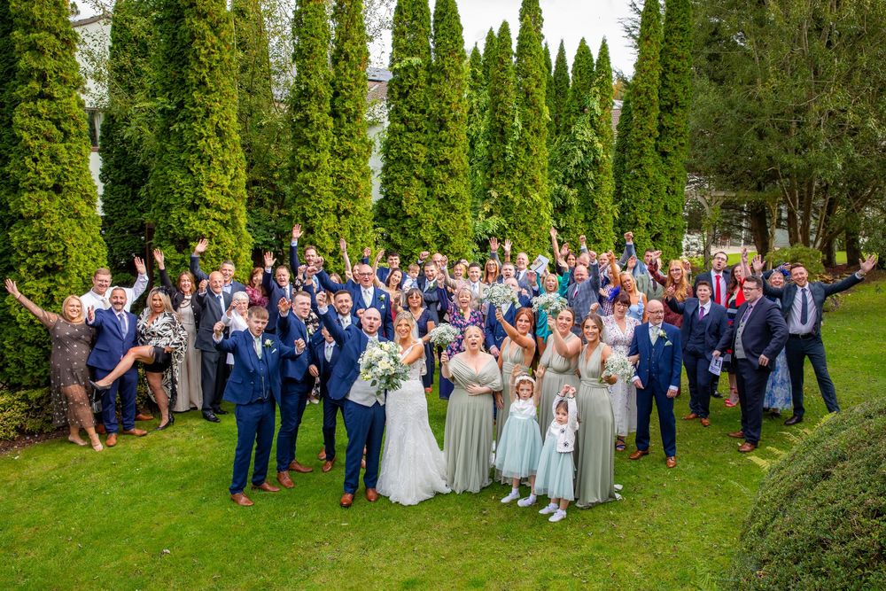 Large wedding party group photo with bride, groom, and guests celebrating outdoors among tall trees.