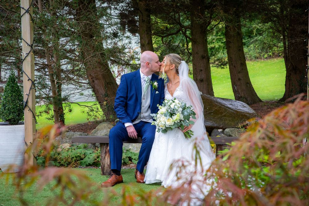 Bride and groom share romantic moment seated among lush greenery with bouquet of white flowers.