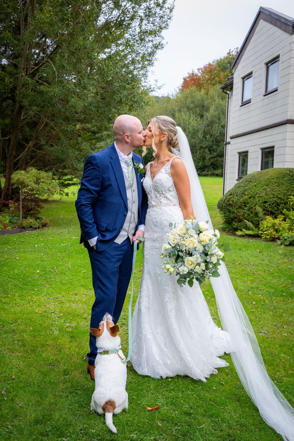 Bride and groom kiss on green lawn with small dog watching, holding white floral bouquet.
