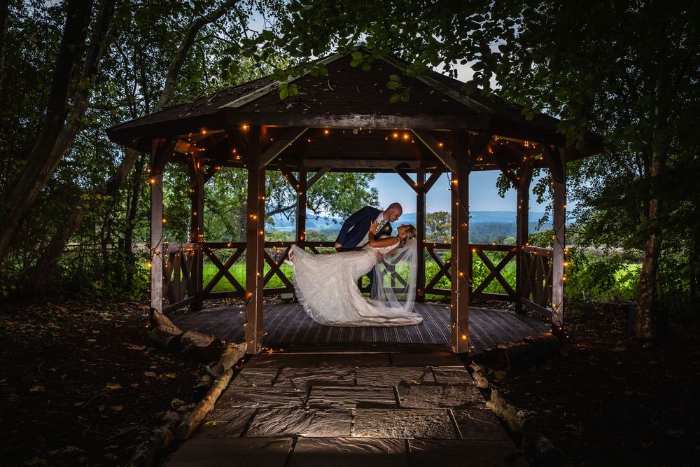 Bride and groom share romantic kiss in fairy-light gazebo overlooking scenic ocean view at dusk.