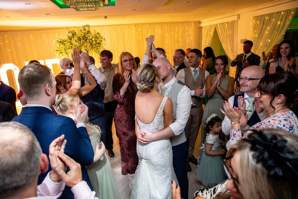Bride and groom share first dance surrounded by cheering guests in elegantly decorated reception hall.