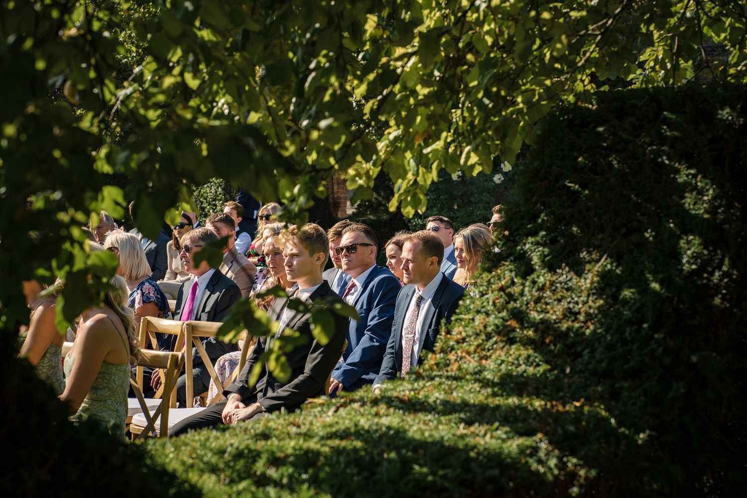 Wedding guests seated outdoors in rows of white chairs surrounded by lush greenery during a garden ceremony.
