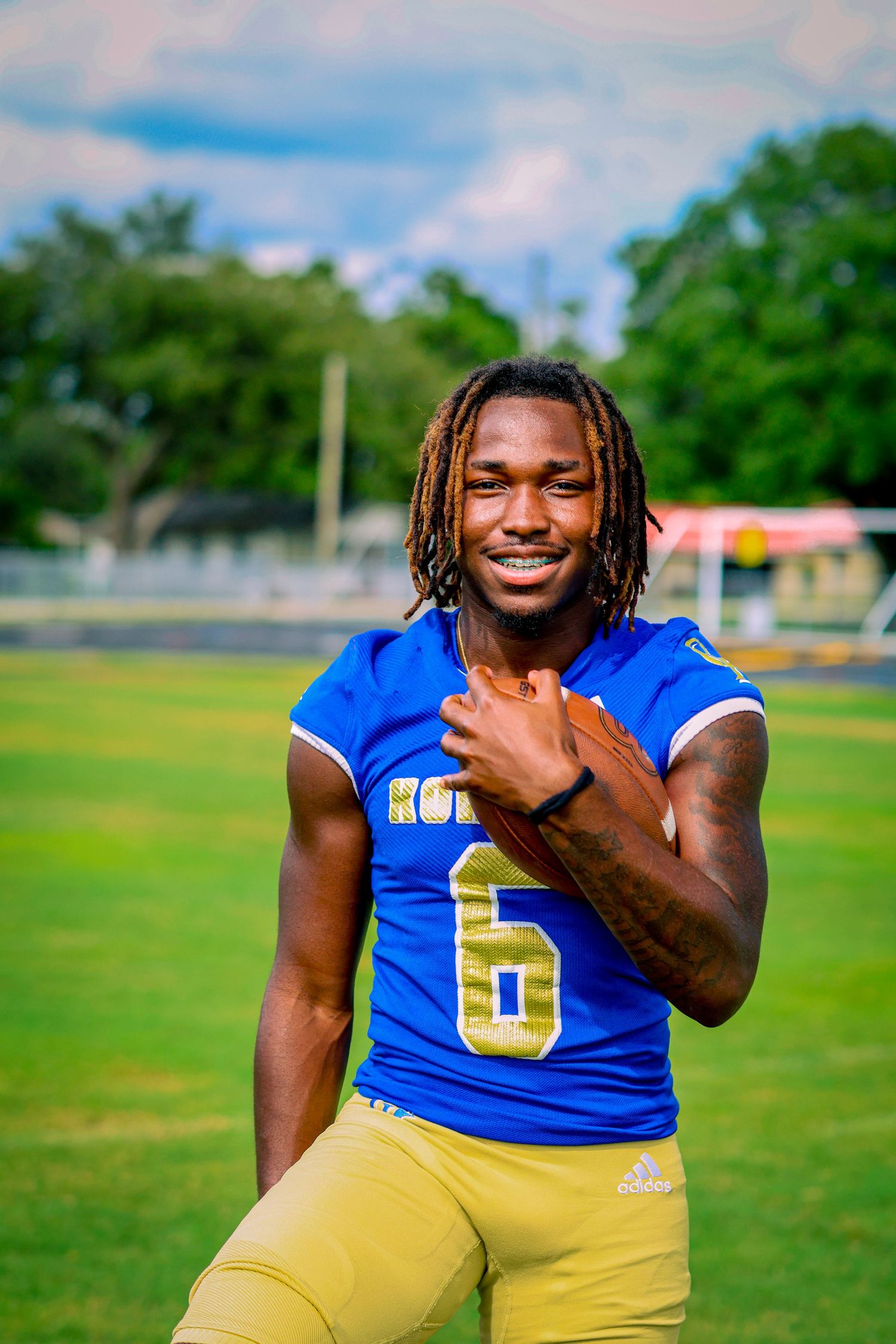 Football player in blue and gold uniform sits on field smiling at camera during sunny outdoor photoshoot.