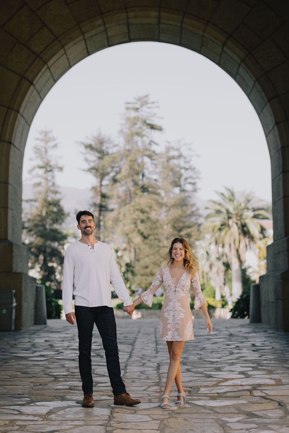 Couple holding hands walking under a stone arch with palm trees in background, woman in lace dress, man in white shirt.
