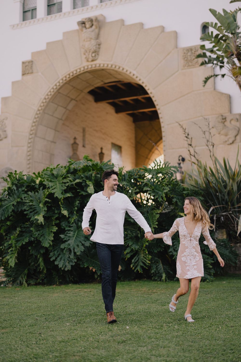A happy couple holds hands and walks on green grass in front of an elegant arched building with lush tropical plants.