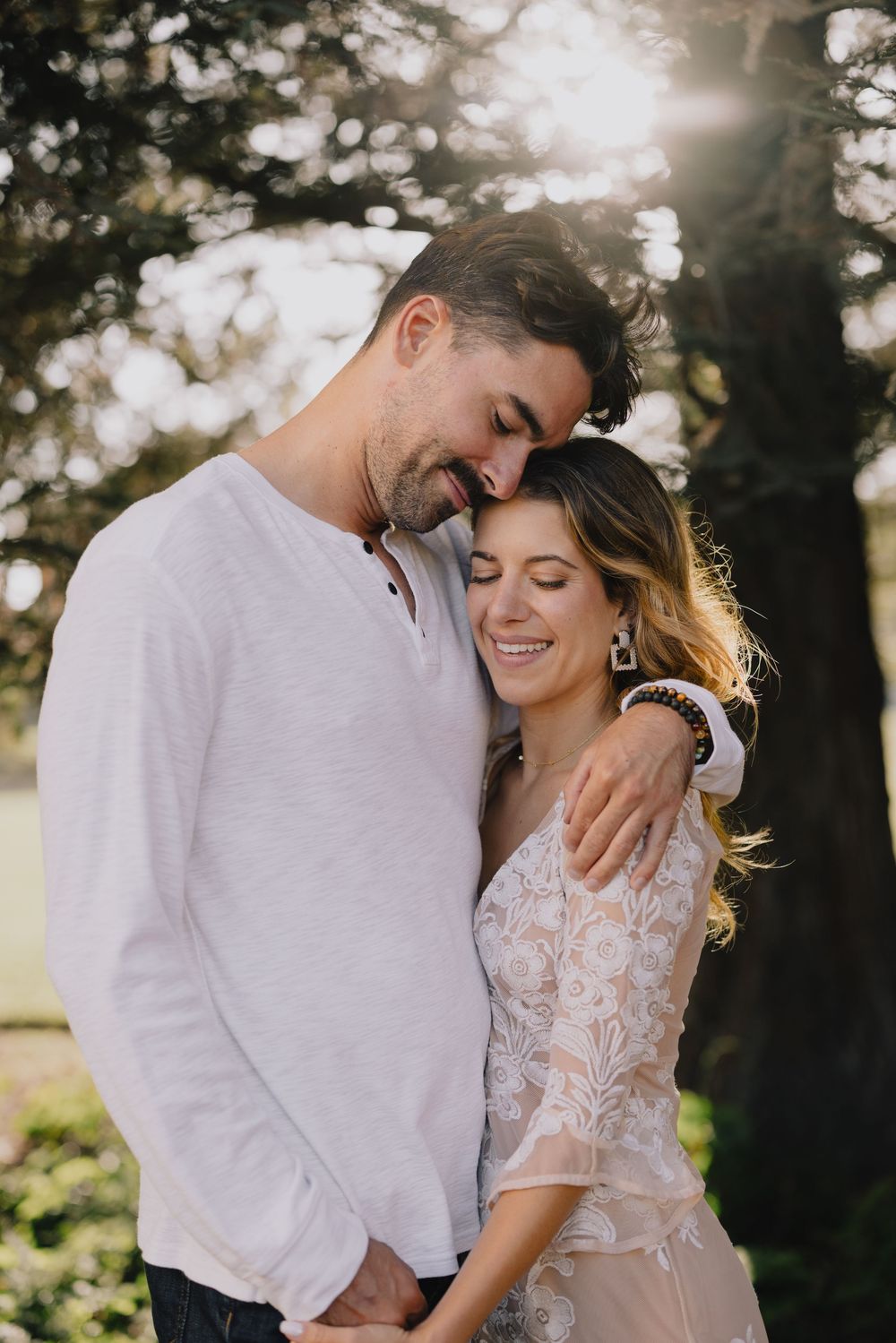 A couple embraces tenderly outdoors, smiling with eyes closed, bathed in warm golden sunlight near a tree.