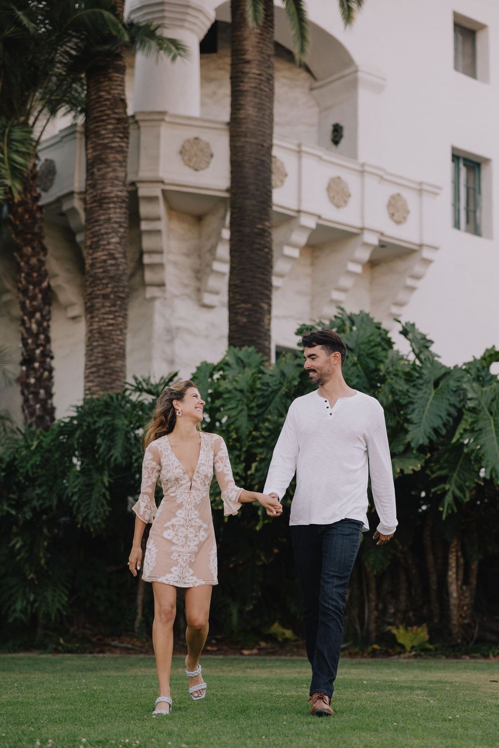 Couple holding hands and walking on green lawn in front of white building with palm trees.