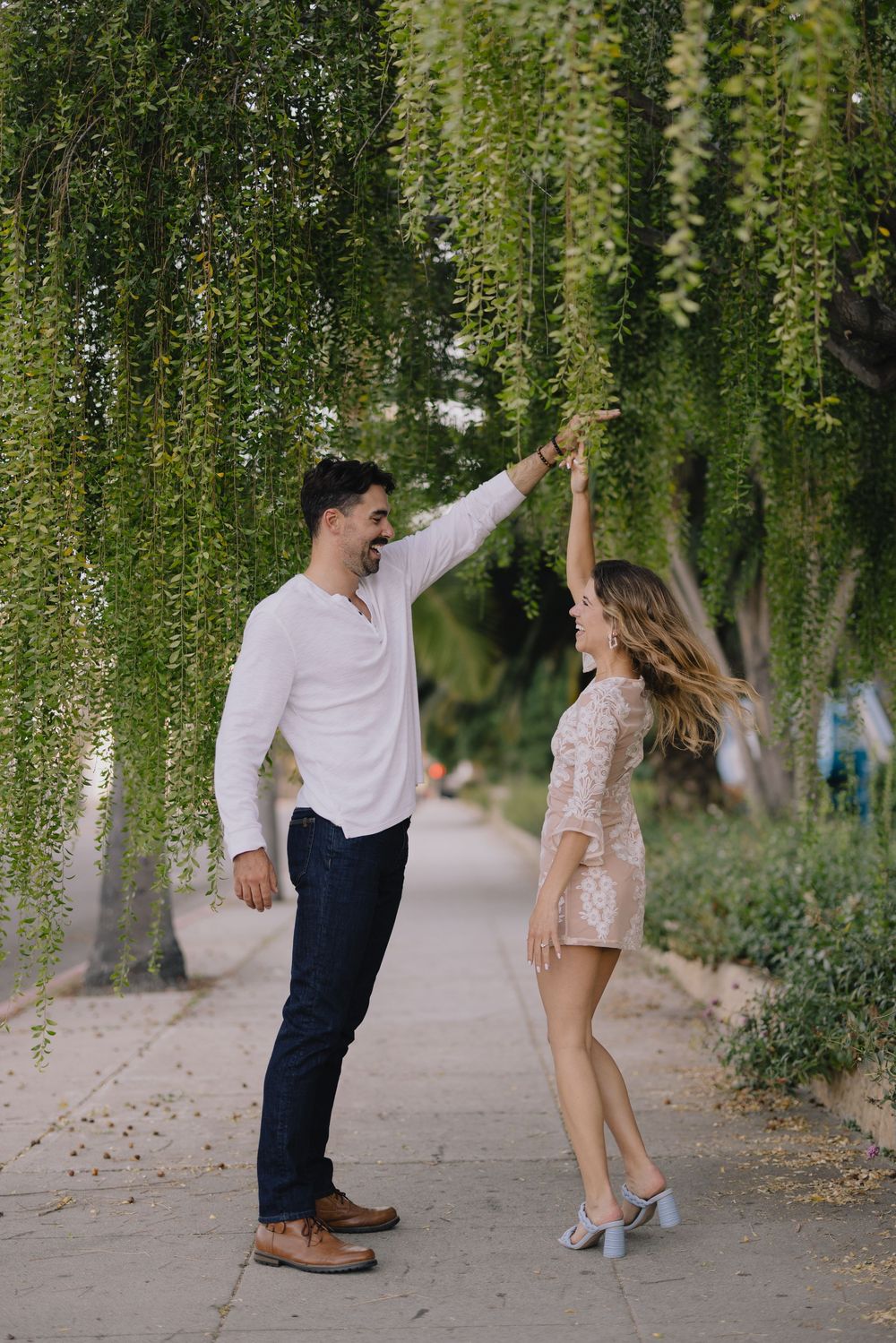 Couple dancing outdoors under weeping willow trees on a pathway, man in white shirt spinning woman in pink lace dress.