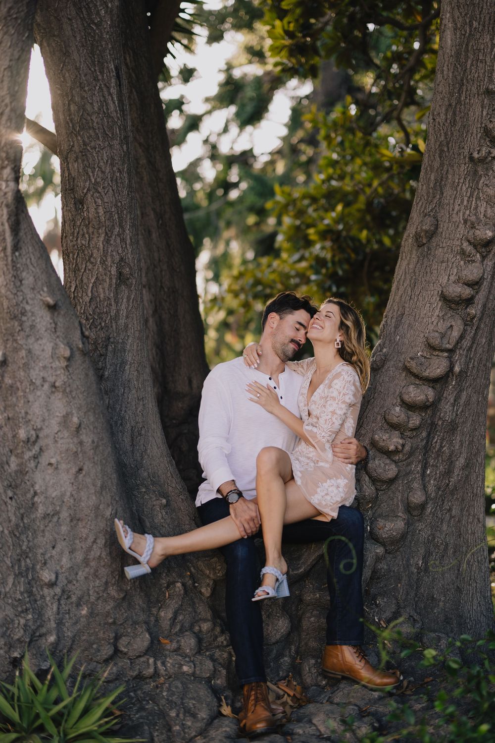 Couple sharing a romantic moment sitting between two large trees in golden sunlight.