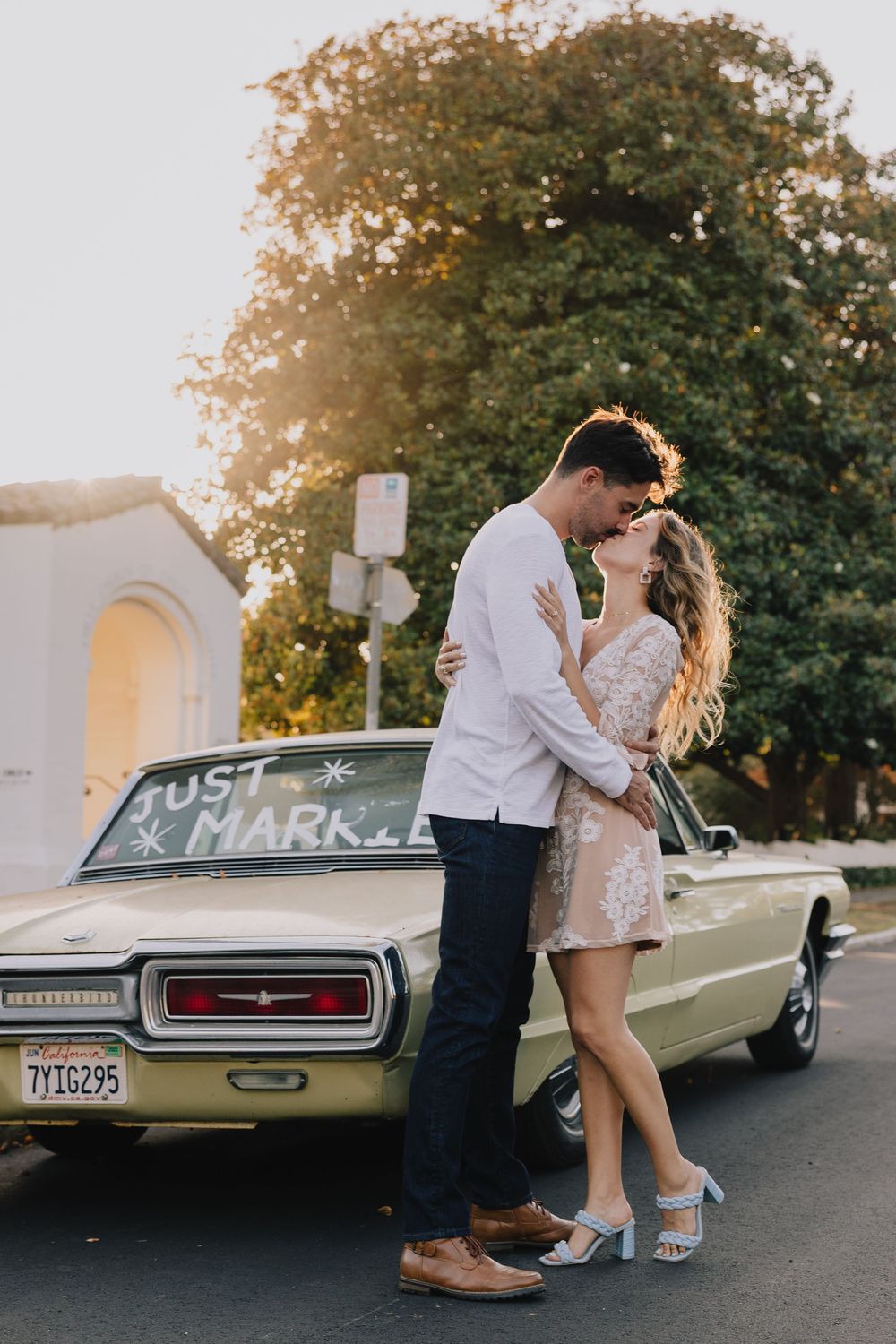 Newlywed couple kissing beside a vintage car with Just Married sign in warm golden sunset light.