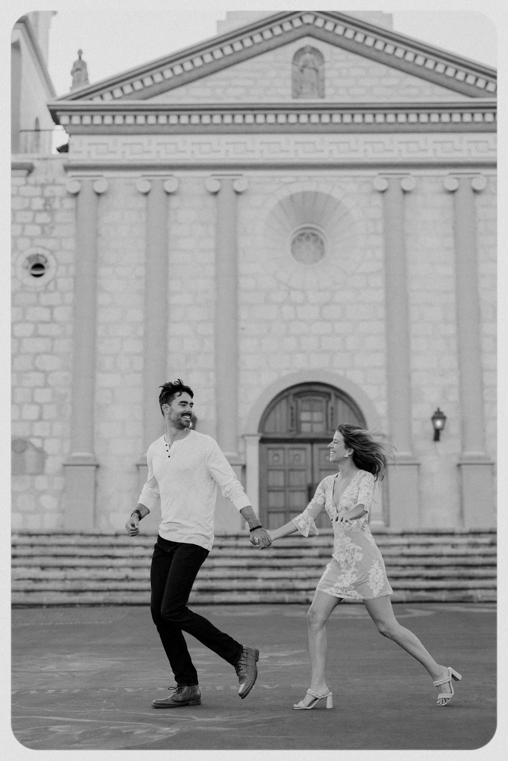 A joyful couple holds hands and walks in front of a pink colonial church building.
