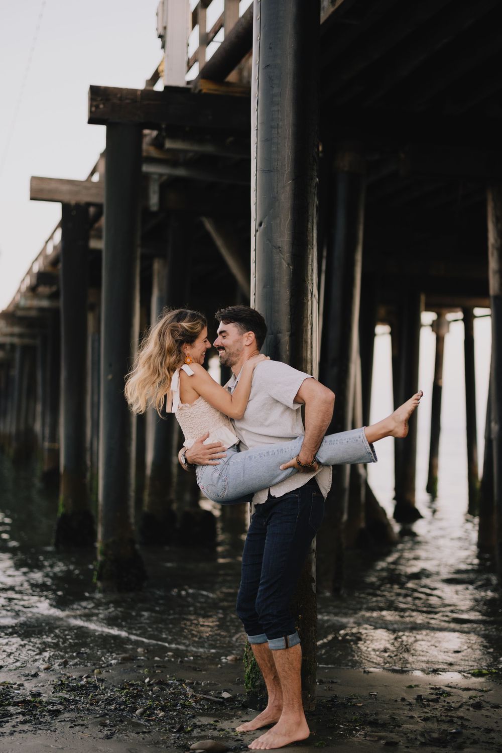 Couple sharing a romantic moment under a wooden pier, man lifting woman while barefoot on wet sand.