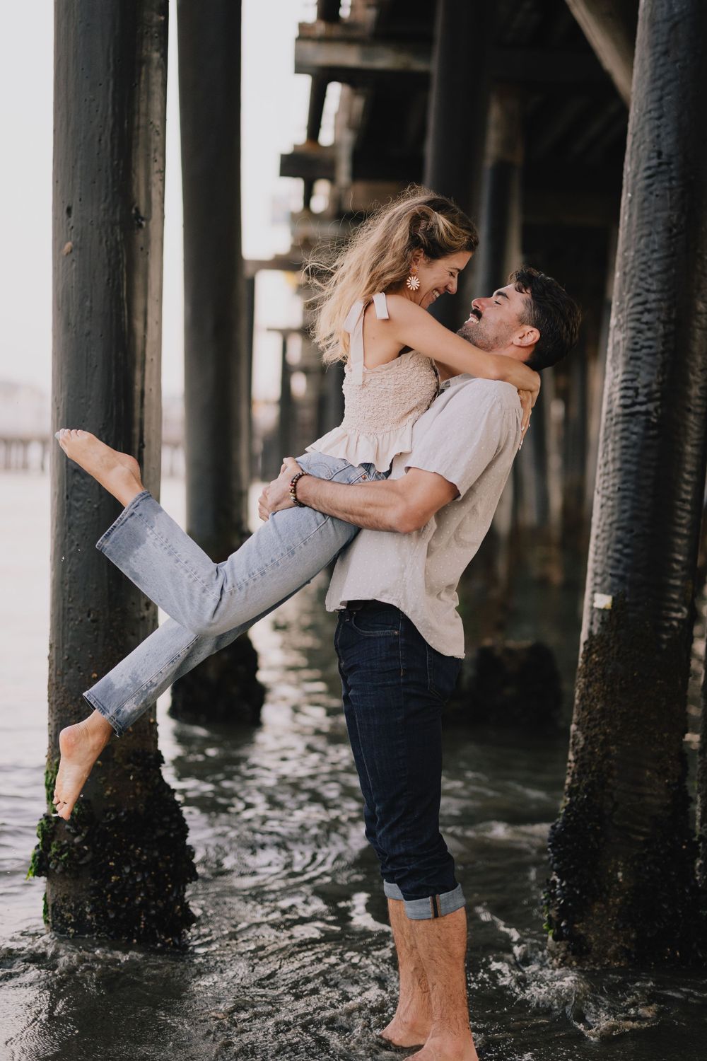 A man lifts a woman joyfully under a beach pier, both barefoot in jeans, sharing a romantic moment in shallow water.