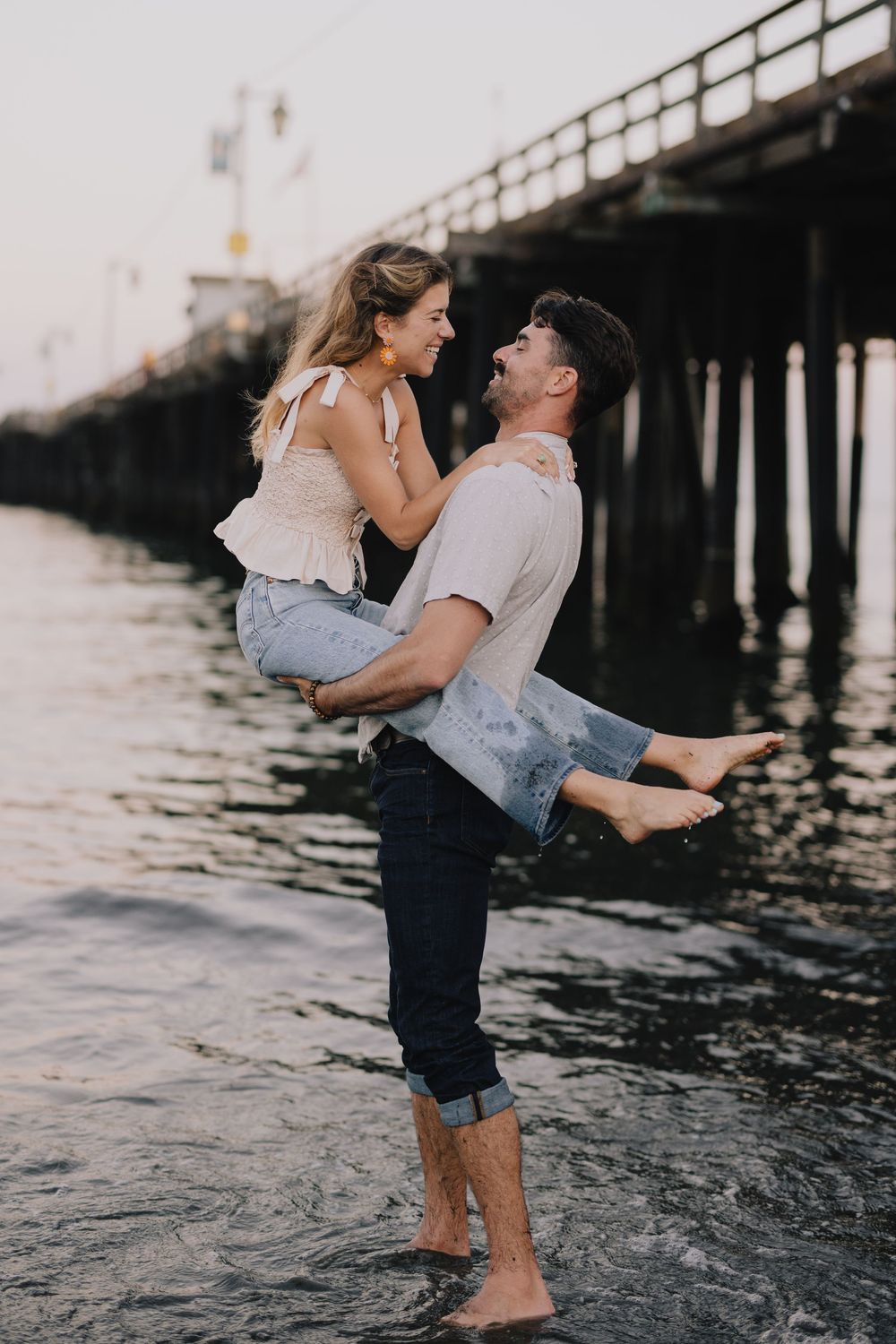 Barefoot couple laughing as man lifts woman near a pier, standing in shallow ocean water at sunset.