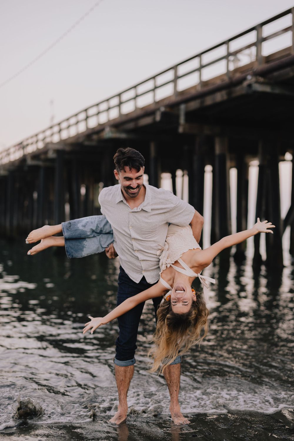 Man joyfully swings laughing woman upside down near a wooden pier over shallow water at sunset.