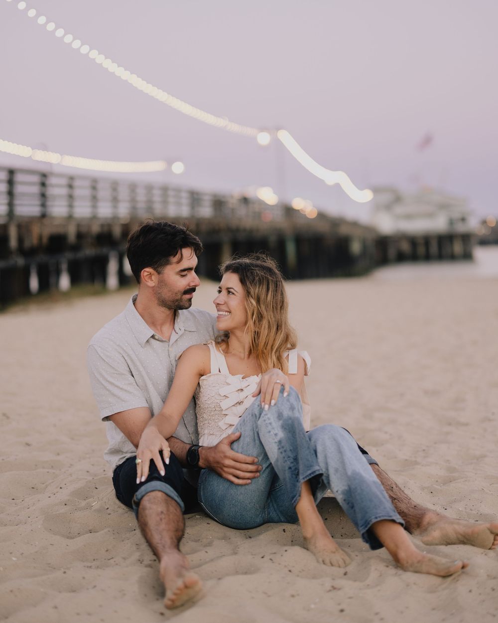 Couple sitting on beach sand at dusk near a pier with string lights, sharing a romantic moment.
