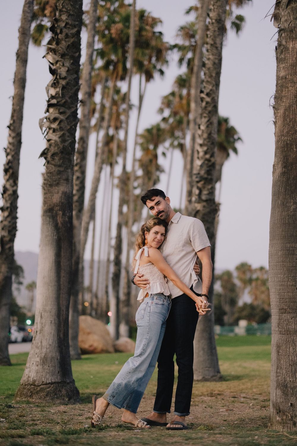 A romantic couple embraces among tall palm trees in a lush green park at dusk.